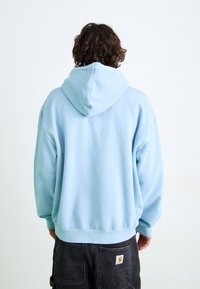 Light blue hoodie with a drawstring hood, ribbed cuffs and hem, made from soft fabric, worn with black pants featuring back pockets.