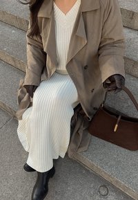 Brauner Trenchcoat über einem weißen Strickkleid, schwarze Stiefeletten und eine braune Schultertasche. Strukturiertes Material und glatte Lederdetails sind sichtbar.