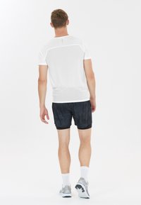 Homme marchant de dos, portant une chemise blanche à manches courtes, un short de sport noir, des chaussettes blanches et des chaussures de course grises sur un fond blanc.