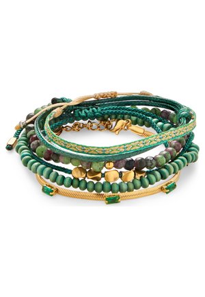 Stak af grønne perle- og flettede armbånd med gulddetaljer og grønne ædelstene, arrangeret i en cirkulær bunke på hvid baggrund.