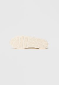 Σόλα παπουτσιού σε μπεζ καουτσούκ με υφή και οριζόντιες αυλακώσεις για πρόσφυση. Στρογγυλή μορφή με το λογότυπο της μάρκας κοντά στη φτέρνα.