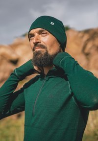 Maglia verde con zip e maniche lunghe realizzata in tessuto strutturato, abbinata a un berretto coordinato. Presenta un piccolo logo e un design aderente.