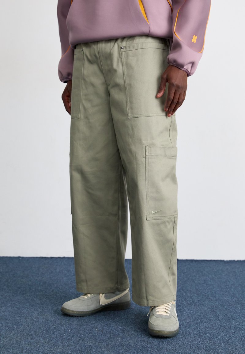 Grønne cargo-bukser med løs passform, flere lommer og en subtil Nike-logo. Materialet virker robust med en matt tekstur.