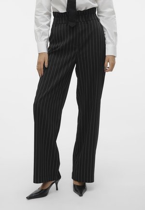 Woman wearing black high-waist, straight-leg pinstripe pants with a white shirt, black tie, and black pointed-toe heels.