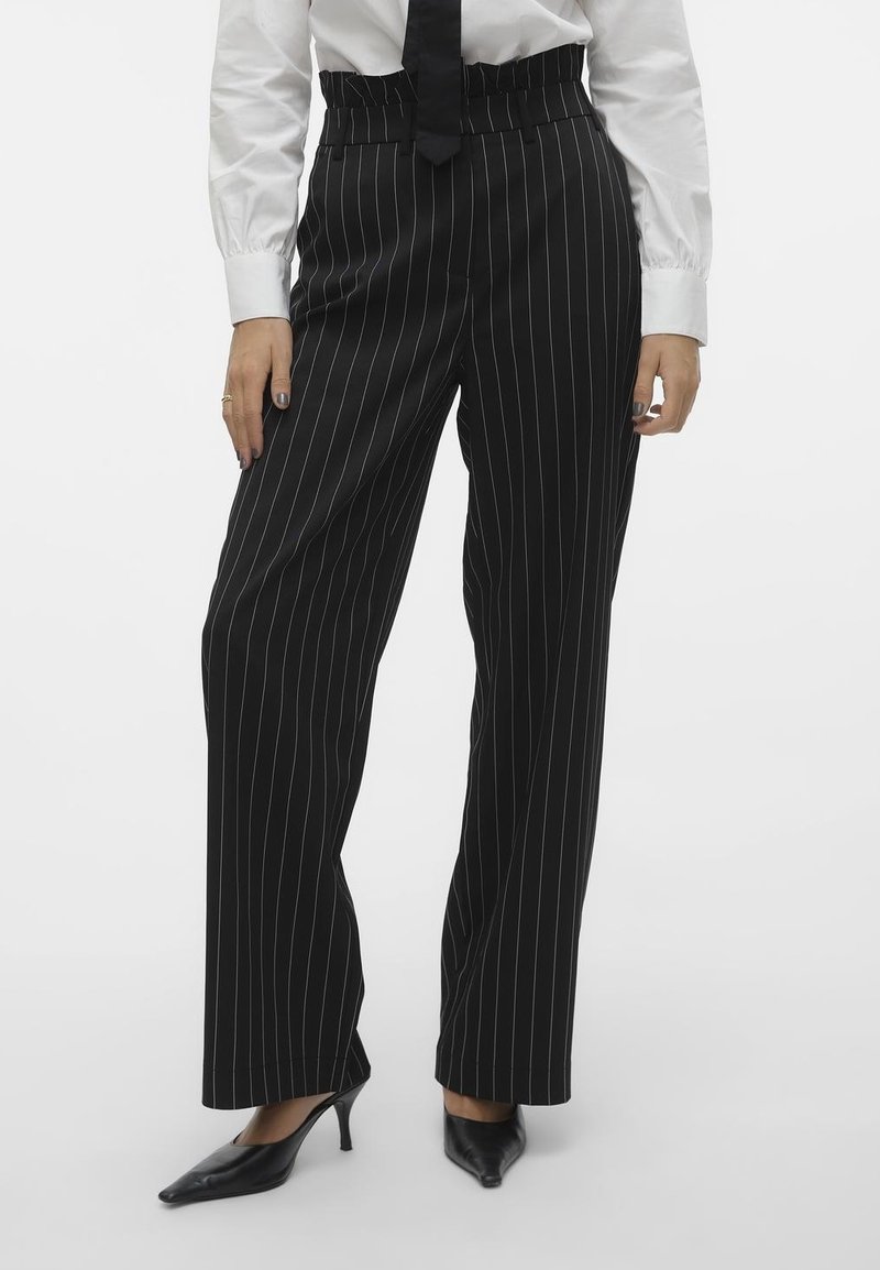 Femme portant un pantalon noir taille haute à jambes droites à rayures fines, avec une chemise blanche, une cravate noire et des talons noirs à bouts pointus.