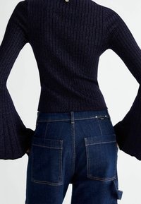 Personne portant un pull côtelé bleu foncé avec des manches évasées et un jean en denim bleu foncé avec de grandes poches arrière, vue de dos.