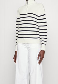 Pullover lavorato a maglia color crema con strisce orizzontali blu navy, colletto alto e texture a coste. Abbinato a pantaloni bianchi a gamba larga.