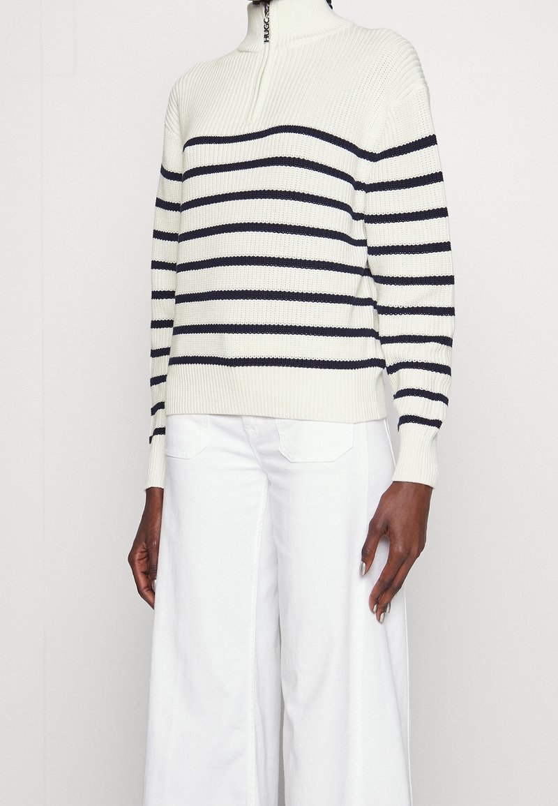Pullover lavorato a maglia color crema con strisce orizzontali blu navy, colletto alto e texture a coste. Abbinato a pantaloni bianchi a gamba larga.