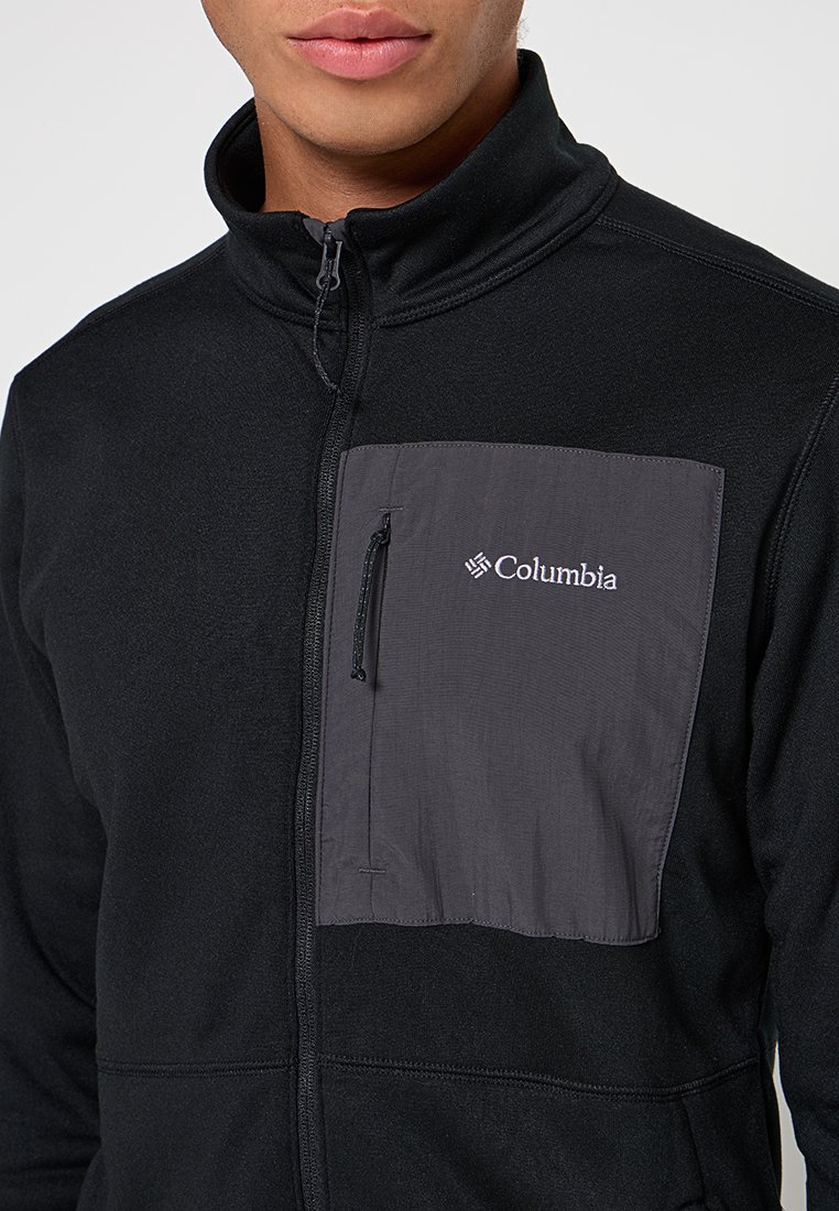Veste en polaire noire avec une poche poitrine zippée grise. Présente un col haut et le logo Columbia sur la poche. Texture lisse.