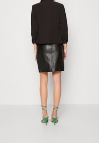 Blazer noir court avec manches froncées, associé à une jupe en cuir noire ajustée et des talons verts à lanières, mettant en valeur un contraste de couleurs vibrant.