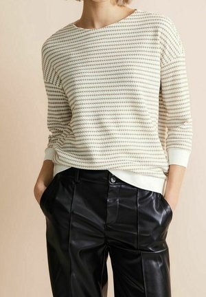 Persoon die een beige-wit gestreepte trui met lange mouwen en zwarte leren broek draagt, met handen in de zakken.