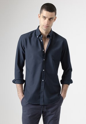 Hombre con las manos en los bolsillos, vistiendo una camisa azul marino oscuro con las mangas remangadas y pantalones a juego, de pie contra un fondo liso y claro.