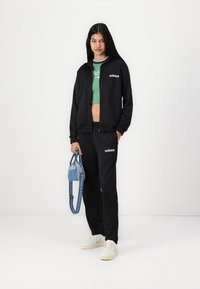 Fekete cipzáras kapucnis felső fehér logóval, zöld crop top, fekete melegítőnadrág és fehér sportcipő. Kék farmertáska van a kezében pánttal.