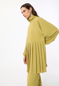Faltige olivgrüne Jacke mit hohem Kragen und langen Ärmeln. Weicher Stoff, mit einem Reißverschluss vorne und strukturierten Falten in der Taille.