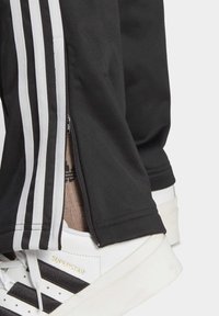 Pogled od blizu na crne trenerke s bijelim prugama i bočnim patentnim zatvaračem, uz bijele Adidas Superstar tenisice s crnim prugama.