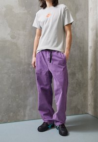 Person standing wearing white Nike t-shirt, loose purple pants with black belt, and black sneakers with teal accents against a concrete wall.