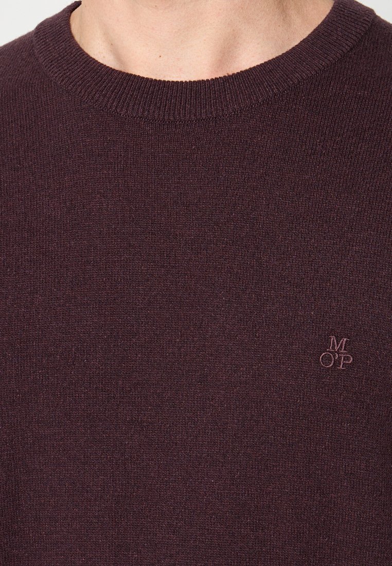Nahaufnahme einer Person, die einen dunkelvioletten Strickpullover mit Rundhalsausschnitt und kleinem gesticktem "M OP"-Logo auf der Brust trägt.