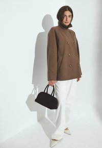 Woman wearing a brown double-breasted coat, white wide-leg pants, white pointed shoes, holding a black handbag against a white wall.