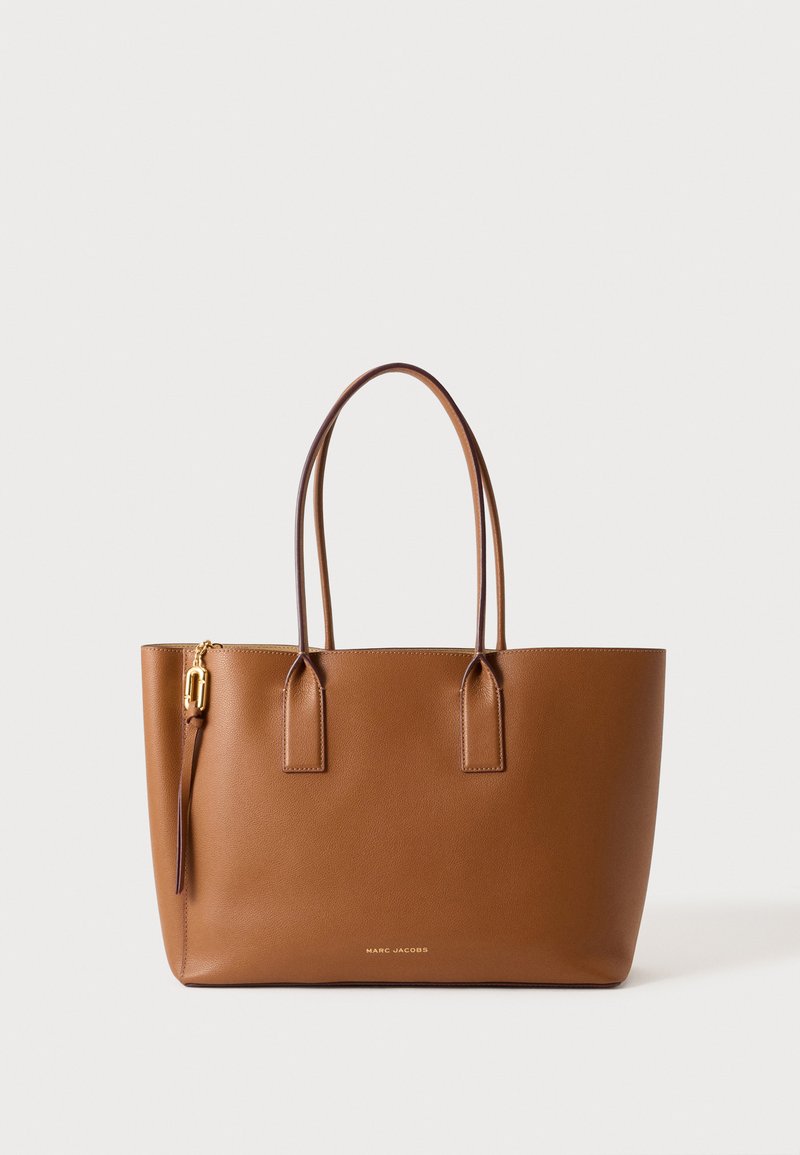 Sac cabas en cuir marron avec deux longues anses et une petite tirette de fermeture éclair dorée sur le côté, sur un fond clair uni.