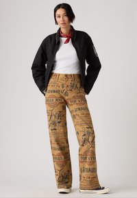 Chaqueta negra con cremallera y tachuelas doradas, top blanco de canalé y pantalones de pierna ancha marrones con gráficos de Toy Story. Zapatillas negras completan el look.