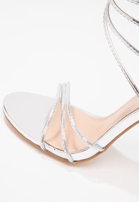 Sandale argentée à brides avec une semelle blanche, présentant de fines brides métalliques et une semelle intérieure beige lisse. Légèrement texturée avec un design élégant.
