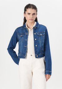 Jaqueta de ganga cortada, azul escura, com botões prateados e dois bolsos no peito, combinada com um suéter de cor clara e calças de tom creme.
