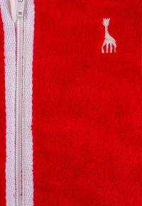 Giacca a zip in pile rosso con zip bianca e un piccolo ricamo di una giraffa beige sull'area del petto. Texture morbida.