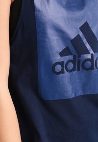 Tummansininen toppi, jossa on kiiltävä sininen suorakulmainen paneeli ja musta "adidas"-logo. Tasainen kankaan pinta, olkapäät paljastava malli.