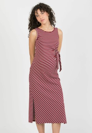 Kobieta o kręconych włosach, ubrana w czerwoną i białą, bezrękawną sukienkę midi w paski, z bocznym rozcięciem i wiązaniem w talii.