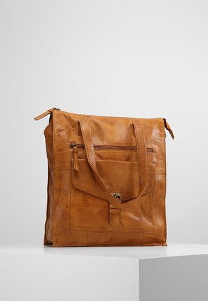 Bruine leren totebag met twee schouderbanden, een voorste ritstasje en een klepzak met koperen gesp, tentoongesteld op een witte ondergrond.