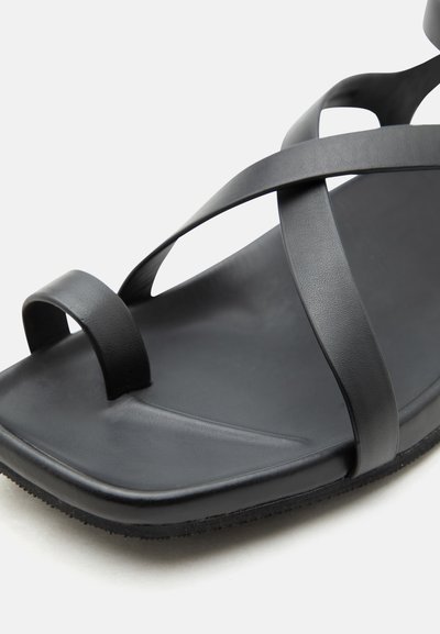 Μαύρο επίπεδο σανδάλι με διασταυρούμενους λεπτούς ιμάντες και θηλιά γύρω από το μεγάλο δάχτυλο, με λείο πέλμα και υφασμένο πέλμα.