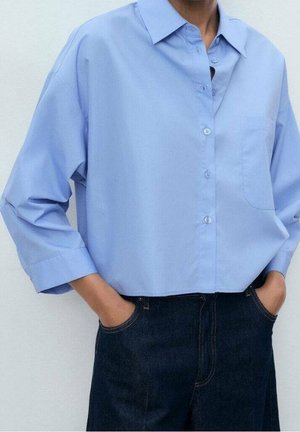 Personne portant une chemise à manches longues bleu clair boutonnée et rentrée dans un jean bleu foncé, les mains dans les poches, sur un fond uni.