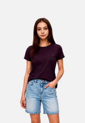 Junge Frau mit langen braunen Haaren, die ein dunkelviolettes T-Shirt und hellblaue Jeansshorts trägt, mit einer Hand in der Tasche.