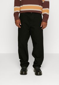Pantalones de algodón negros con un ajuste relajado, con bolsillos frontales y pasadores para cinturón, combinados con zapatos de cuero negros y una camisa de manga larga a rayas en color burdeos.