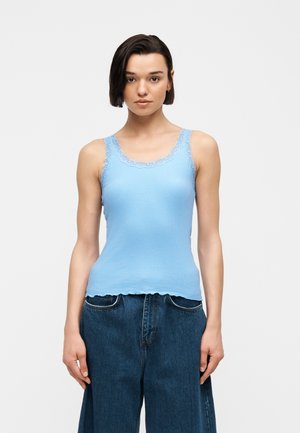 Ung kvinde med kort mørkt hår iført en lyseblå top med blonderkant og løstsiddende mørkeblå jeans, stående mod en ensfarvet baggrund.