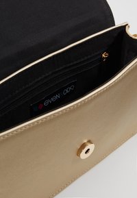Guldig metallisk clutchväska med svart tygfoder, innehållande en ficka med dragkedja, tryckknappsstängning och en slät yta.