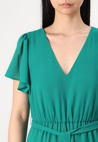 Robe verte avec un décolleté en V profond, des manches flottantes et une taille élastique avec une ceinture assortie. Tissu lisse avec une texture fluide.
