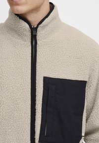 Beige fleece jacket with a high collar, featuring a contrasting black front pocket and zip closure. Soft texture, relaxed fit.