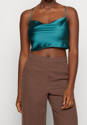 Femme portant un top court en satin bleu sarcelle et un pantalon marron taille haute, debout devant un fond uni.