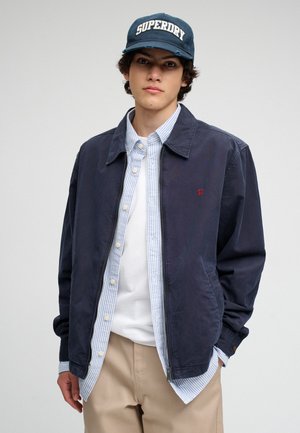 Joven vestido con chaqueta azul marino, camisa de rayas azules y blancas, camiseta blanca, pantalones beige y gorra azul marino con texto "SUPERDRY".