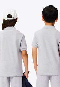 Två barn som bär matchande ljusgrå pikétröjor med korta ärmar och kragar. En av dem håller en mörk jacka. Vit keps på det ena barnet.