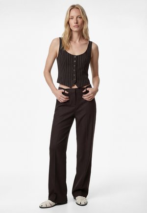 Blonde Frau, die eine schwarze Nadelstreifen-Weste mit Knöpfen, hoch taillierte schwarze Hosen mit weitem Bein und weiße geschlossene Sandalen trägt, steht mit den Händen in der Hüfte.