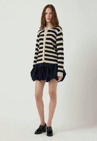 Cardigã riscado em preto e creme com fecho de botão, combinado com uma saia puffy azul escura e mocassins pretos. Usado por uma modelo num ambiente neutro.
