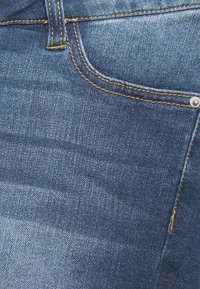 Närbild av en jeansficka i blå denim med gul söm och en metallnitar på höger sida.