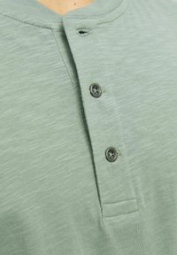 Camisa henley verde claro hecha de una tela suave, con cuello redondo, tapeta de tres botones y un sutil patrón texturizado.