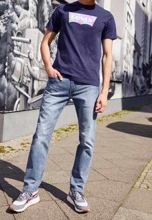 Homme portant un t-shirt Levi's bleu, un jean bleu clair et des baskets grises, debout sur un trottoir avec des murs couverts de fresques en arrière-plan.
