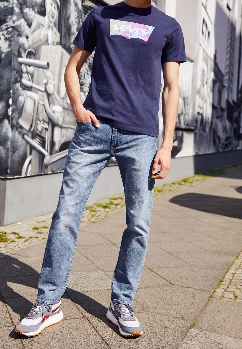 Homme portant un t-shirt Levi's bleu, un jean bleu clair et des baskets grises, debout sur un trottoir avec des murs couverts de fresques en arrière-plan.
