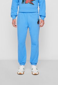 Man wearing light blue sweatpants with black and red embroidered design and white sneakers on a plain gray background.