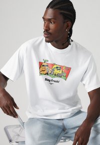 T-shirt bianco a maniche corte con una grafica colorata di frutti antropomorfi con occhiali da sole, testo "Rimani Fresco!" in nero sotto.