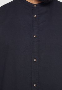 Camicia blu navy con colletto rotondo, cinque bottoni marroni sul davanti e un tessuto morbido e texture.